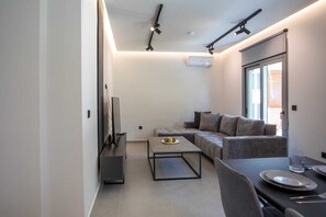 A modern living room with a sectional sofa, a coffee table, a flat-screen TV, and a dining area with a table set for two. - Kennedy 16 Urban Escape Rhodes Town, Apartment (Rhodes)
