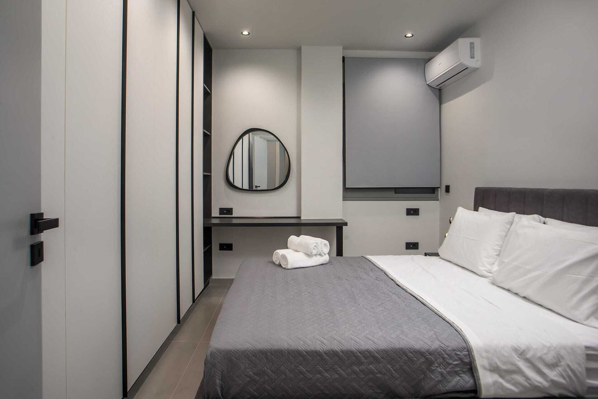 A modern hotel room with a bed, a bedside table with towels, a mirror, and an air conditioning unit.