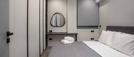 A modern hotel room with a bed, a bedside table with towels, a mirror, and an air conditioning unit.