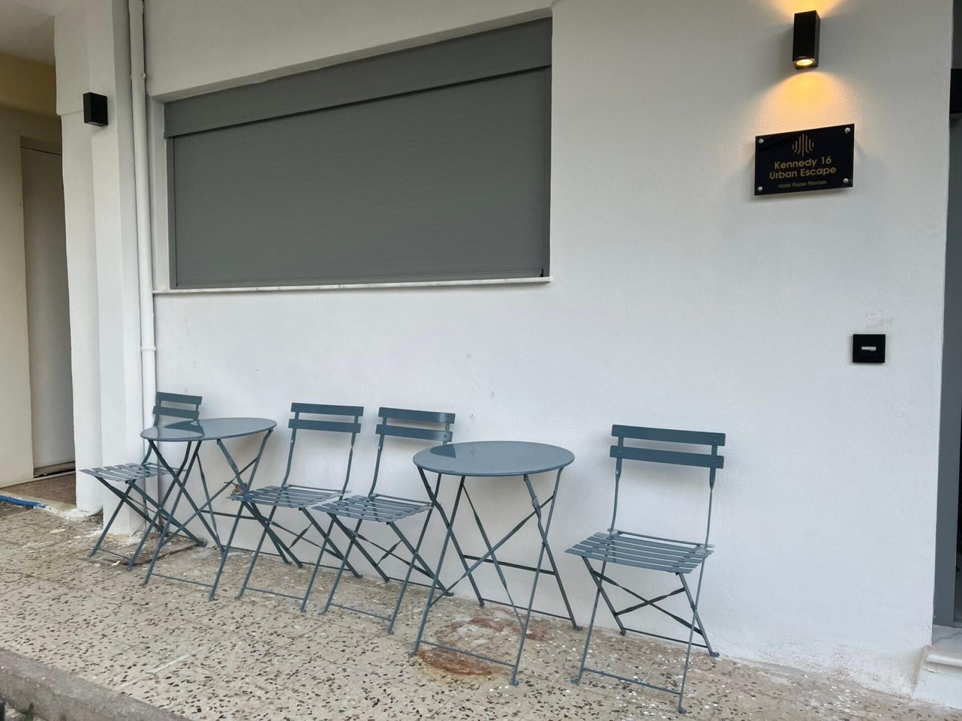Quattro tavoli e sedie all'aperto davanti a un muro bianco, con un cartello che indica 'Kennedy 16 Urban Escape Hotel Aumento Affitto'.