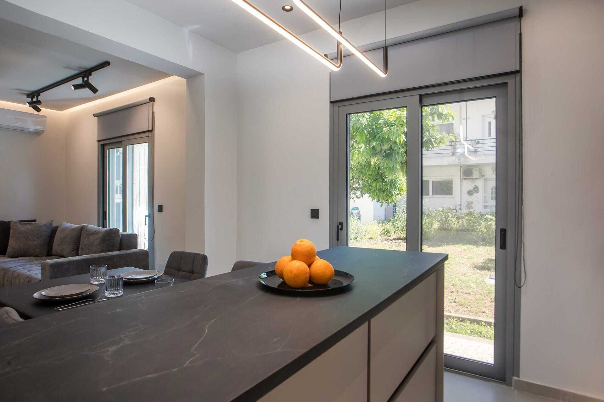 Une cuisine moderne avec un plan de travail foncé, un coin repas avec un canapé et des chaises, et une vue sur l’extérieur grâce à une porte-fenêtre coulissante qui donne sur un espace extérieur.