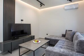 A modern living room with a flat-screen TV, a grey sofa, and a coffee table with fruit. - Kennedy 16 Urban Escape Rhodes Town, Apartment (Rhodes)