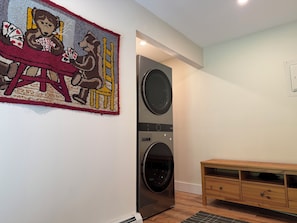 A laundry room with a wall-mounted washer and dryer, a wooden bench, and a rug with a bear and chair design. - Bucks Lodge - Heart of the Catskills (Elka Park)