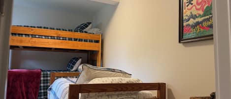 Une chambre avec un lit superposé, un lit simple, un tableau encadré au mur et un panier en osier posé sur une table d’appoint.