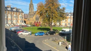 A view from a window overlooking a street with parked cars, a church with a tall spire, and a tree with autumn leaves. - Coastal Place (Scarborough)