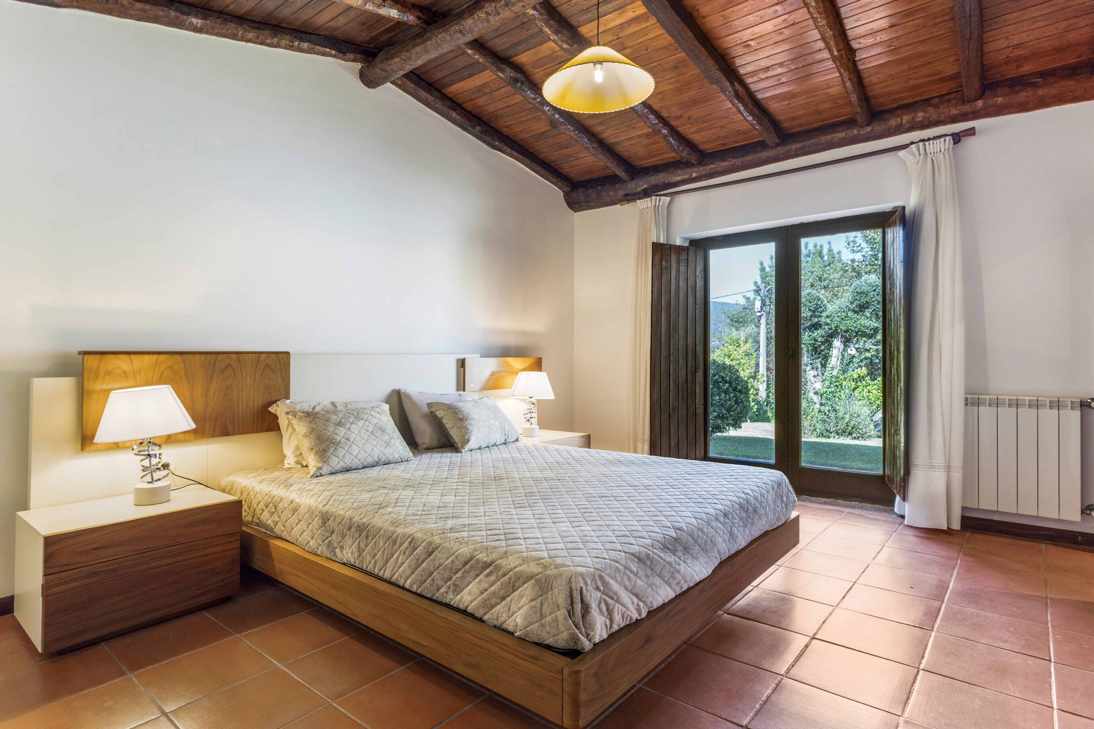 A bedroom with a wooden ceiling, a bed with a quilted bedspread, two bedside lamps, and a view through a window.