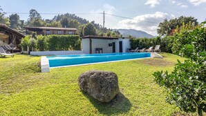 Una piscina con una piedra en primer plano, rodeada de un césped cuidado y un pequeño edificio con fachada blanca.