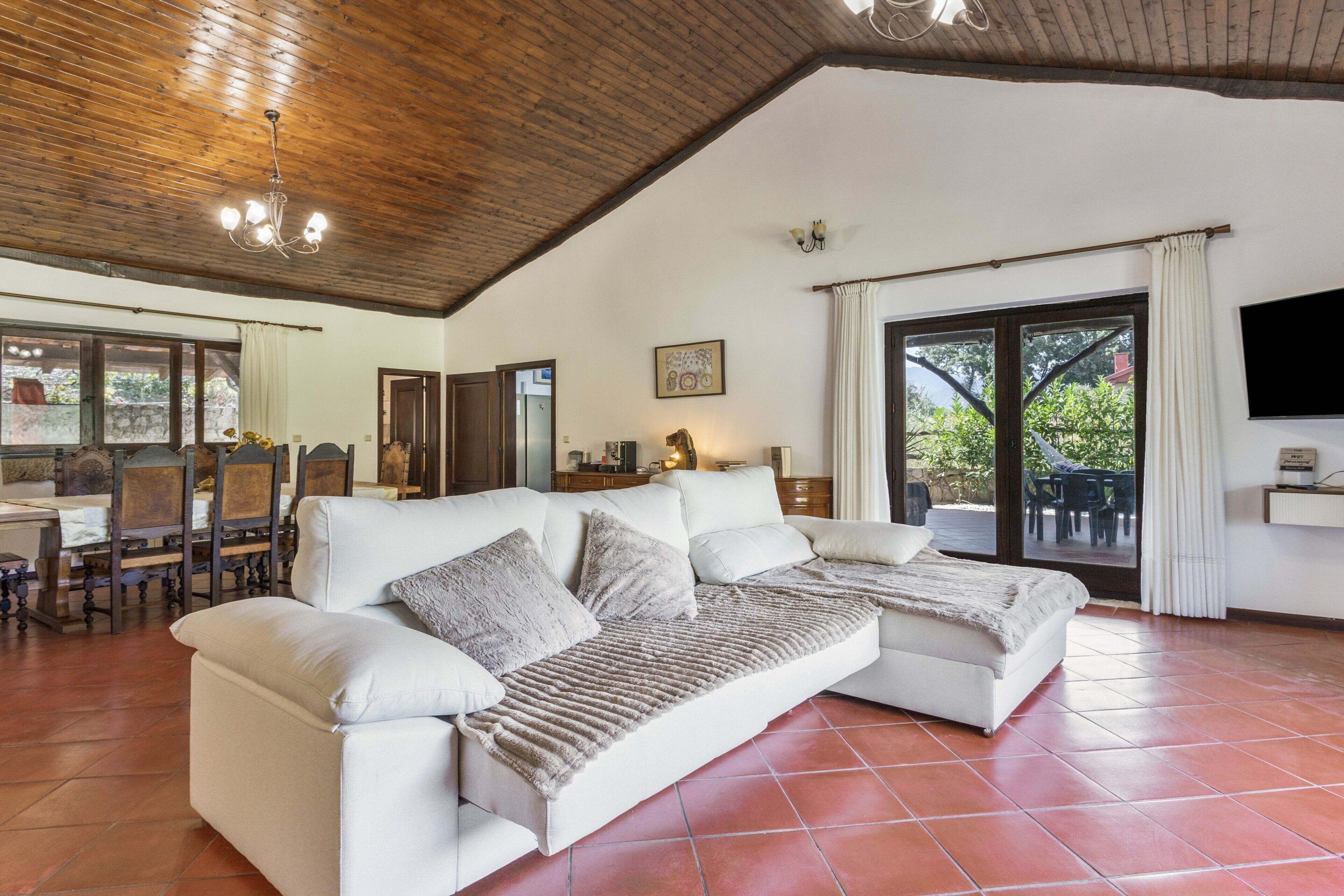 A spacious living area with a large white sectional sofa, a wooden ceiling, and a view through a glass door to an outdoor patio.