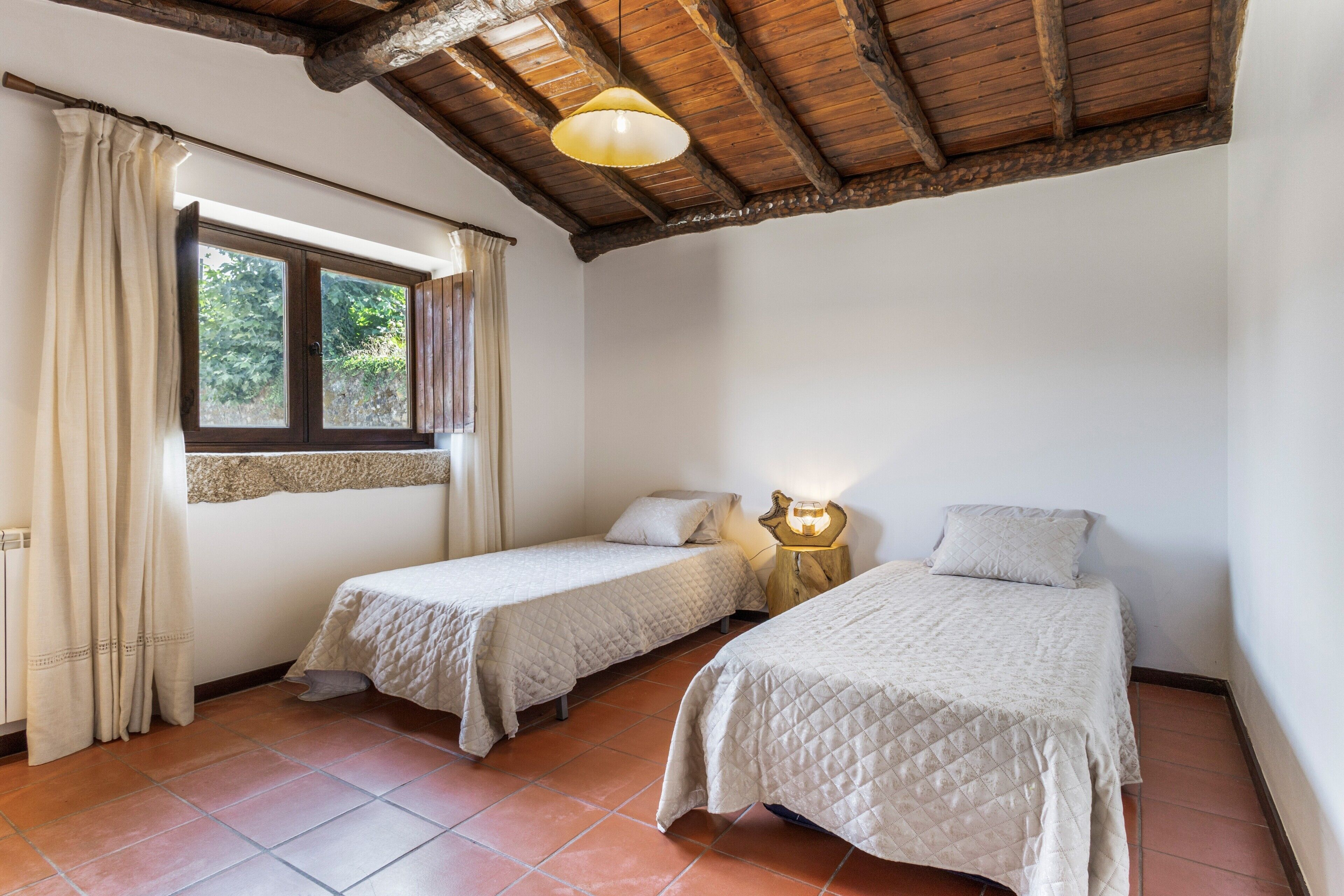 A room with two single beds, a window with curtains, a wooden ceiling, and a small table with a lamp.