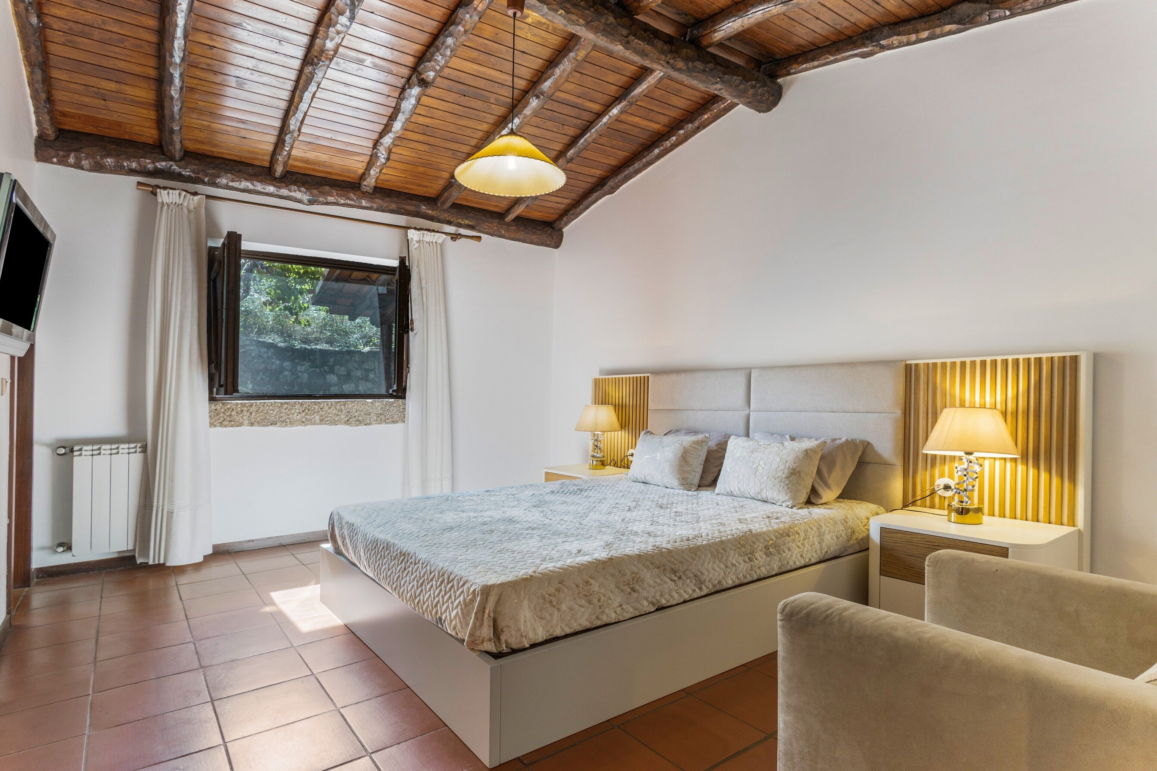 Un dormitorio con cama, dos mesitas de noche con lámparas, una ventana con cortinas y un televisor en la pared.