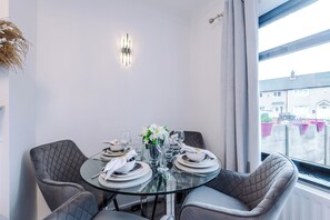 A dining area with a round glass table, grey upholstered chairs, and a white wall with a modern light fixture. - Long Stays | 2 Bed | St Helens | Sleeps 5 (St Helens)