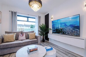 A modern living room with a sofa, a coffee table, a large TV, and a chandelier. - Long Stays | 2 Bed | St Helens | Sleeps 5 (St Helens)