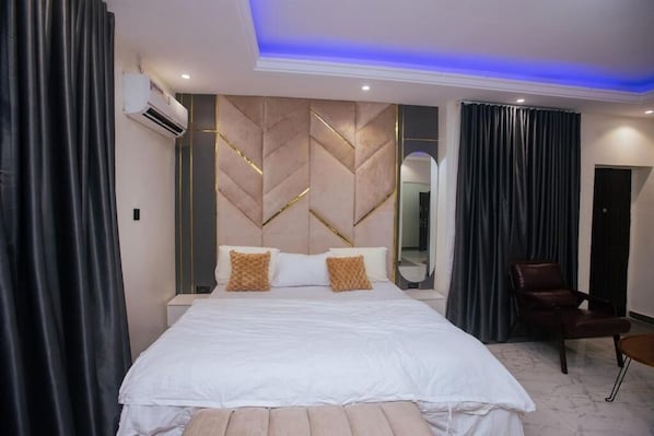 A modern hotel room with a large bed, a headboard with geometric patterns, a seating area with a chair, and a mirror on the wall.