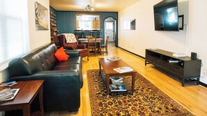 Ein Wohnzimmer mit Sofa, Couchtisch und Fernseher. Im Hintergrund ist eine Essecke mit Stühlen und einem Tisch zu sehen.
