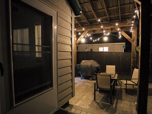 Une terrasse extérieure couverte, agrémentée de guirlandes lumineuses, d’un barbecue abrité et d’une table avec des chaises.