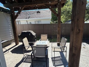 Een overdekt terras met een tafel, stoelen, een barbecue en een houten omheining op de achtergrond.