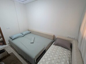 A small, single-bed room with a plain white wall, a wooden headboard, and a patterned bedspread.