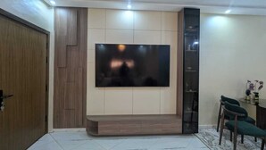 Een woonkamer met een flatscreen televisie aan een wand, een houten deur, een eethoek met stoelen en een betegelde vloer.