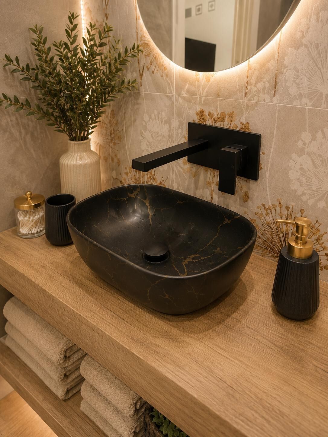 Un baño moderno con un lavabo negro tipo recipiente, un espejo redondo y un florero con flores secas.