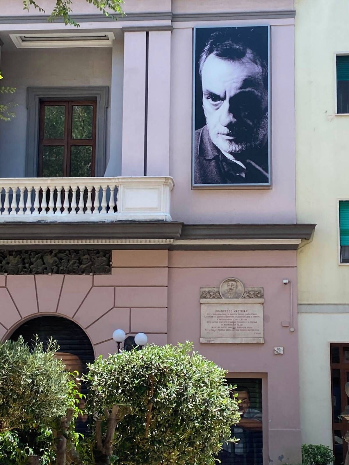 A building facade with a large portrait, a plaque, and a balcony with balustrade.
