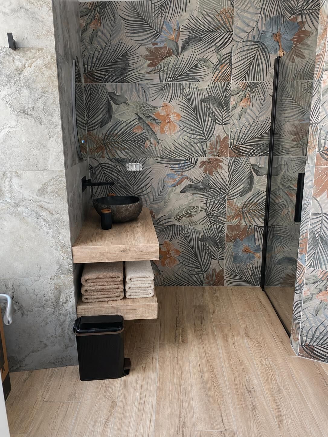 Une salle de bain avec une étagère en bois où sont posées des serviettes, une poubelle noire et un espace douche doté d’une porte en verre.