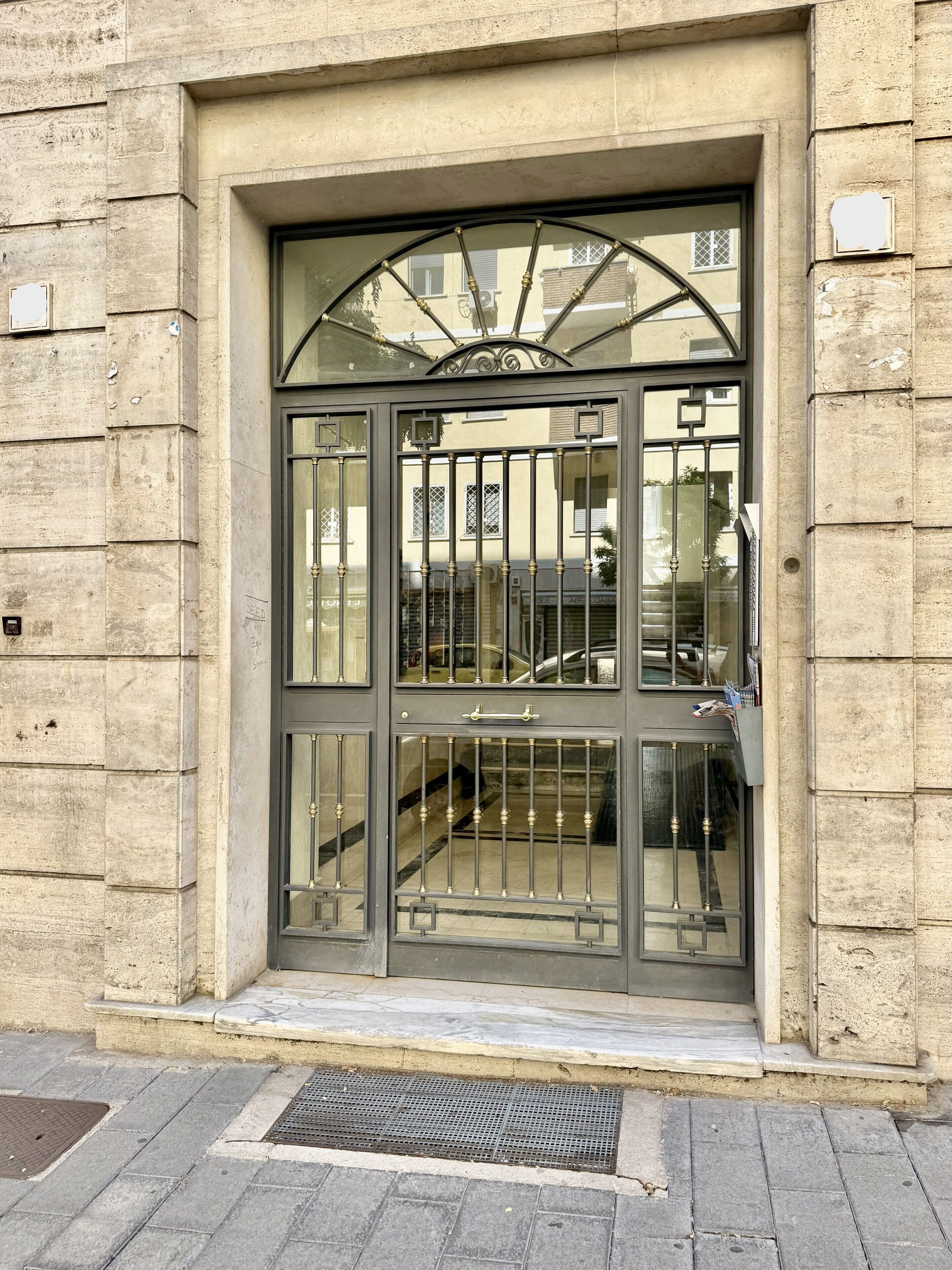 Une entrée d’immeuble avec un portail en métal, une petite marche menant à la porte, et un couvercle de regard sur le sol.