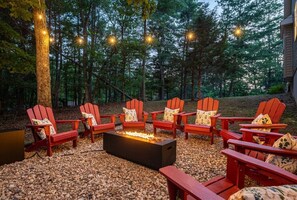Ein Lagerfeuer, umringt von roten Adirondack-Stühlen mit Kissen, auf einem Kiesgrundstück in bewaldeter Landschaft.