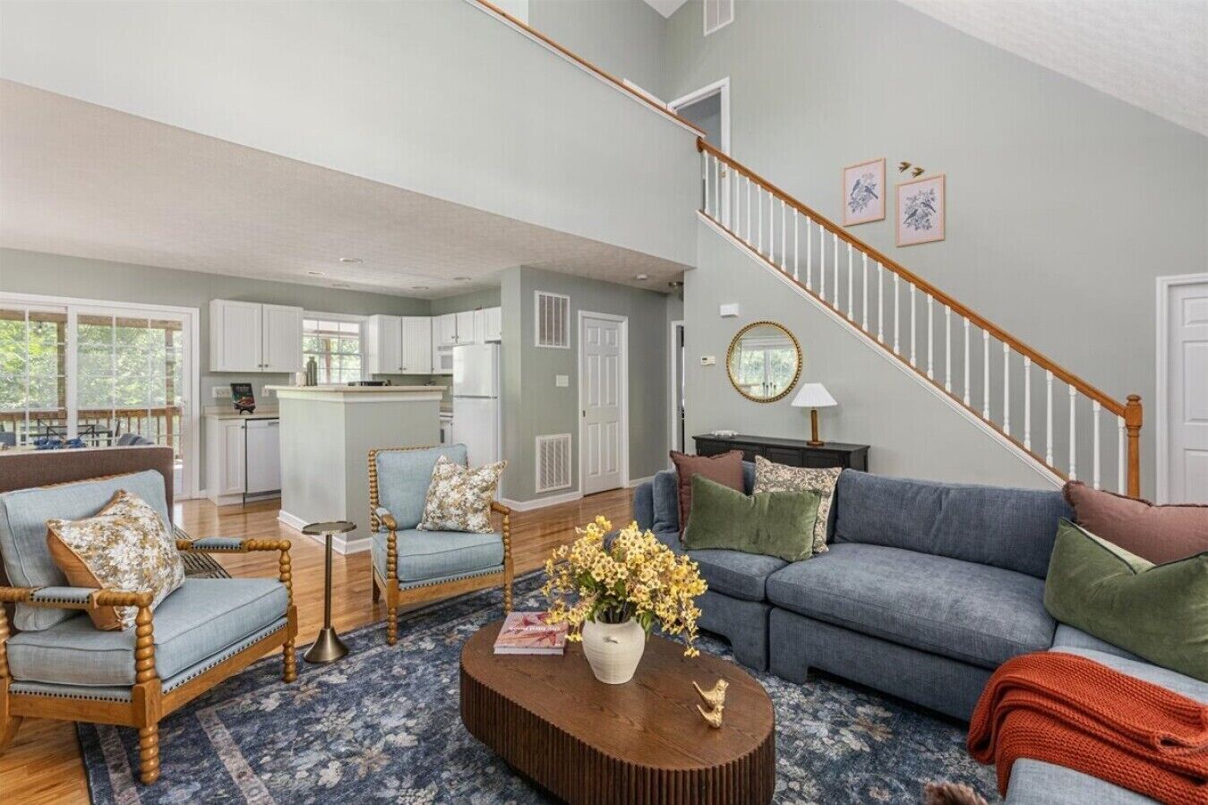 A spacious living room with a blue sofa, a wooden chair, and a coffee table with flowers. A staircase with a wooden railing leads to an upper level.