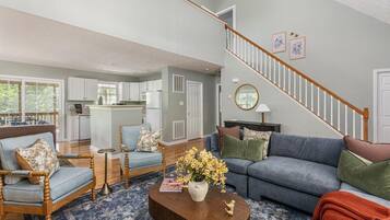 A spacious living room with a blue sofa, a wooden chair, and a coffee table with flowers. A staircase with a wooden railing leads to an upper level.