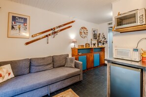 A living room with a sofa, a kitchen area with a microwave, a wooden cabinet, and a wall-mounted clock. - Les Ambrunes - Station de ski accessible à pied (Châtel)