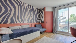 Een slaapkamer met een zwart-witte streep patroon op de muur, een bed met kussens, een stoel en uitzicht op gebouwen door een groot raam.