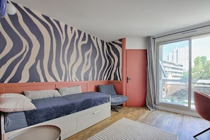 Ein Schlafzimmer mit zebra-gestreifter Wand, einem Bett mit Kissen, einem Sessel und Blick auf Gebäude durch ein großes Fenster.
