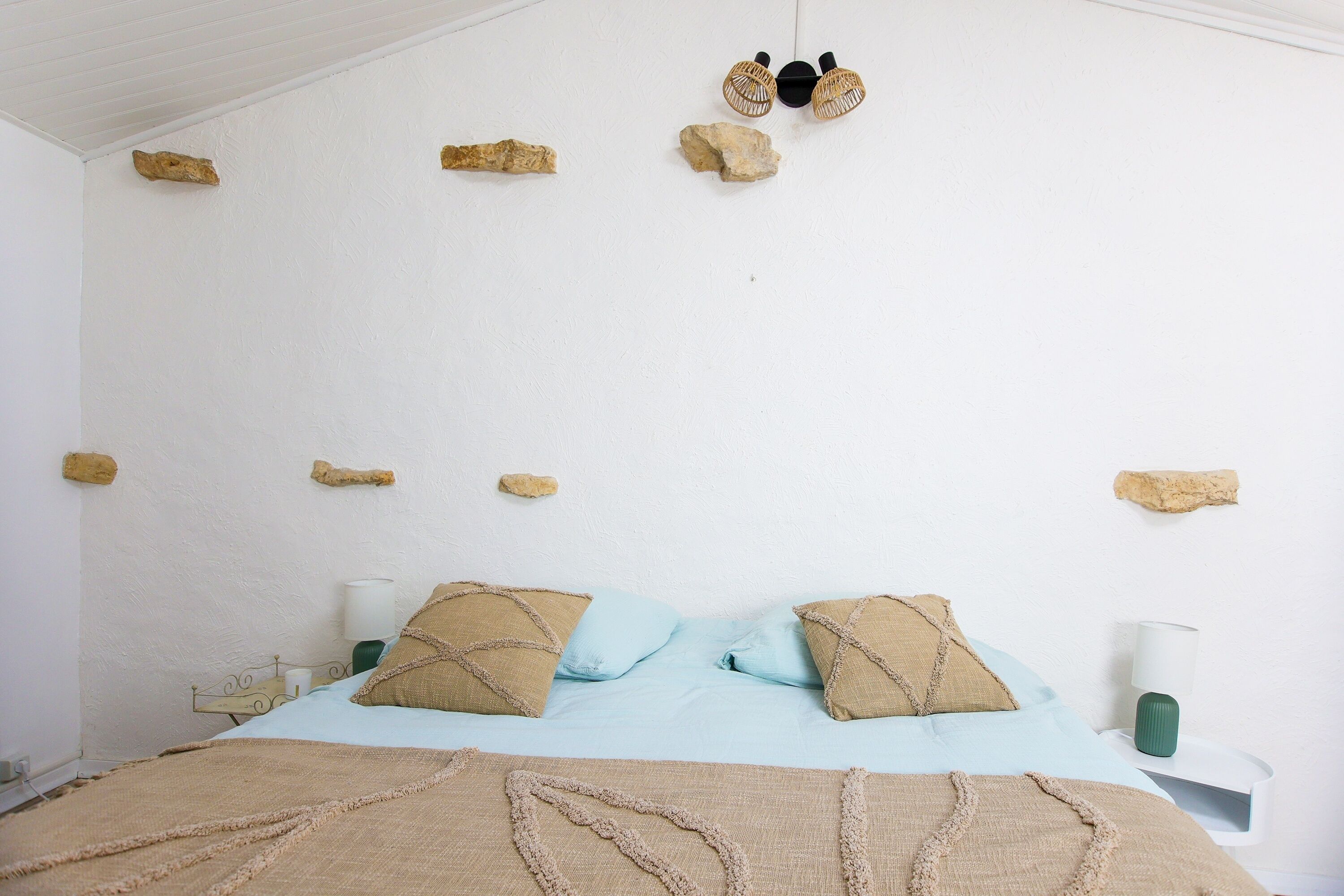 A bed with a light blue bedspread and beige pillows, a white wall with hanging wooden objects, and a bedside table with a green vase.