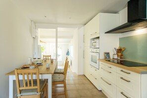 A modern kitchen with white cabinets, a wooden dining table, and a view into the dining area. - Villa Trafal - Proche plage (Saint-Michel-en-l'Herm)