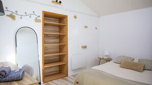 Ein Schlafzimmer mit einem Bett, einem Holzschrank, einem Spiegel, einem kleinen Nachttisch mit Lampe und einer Wand mit einer Leuchte.