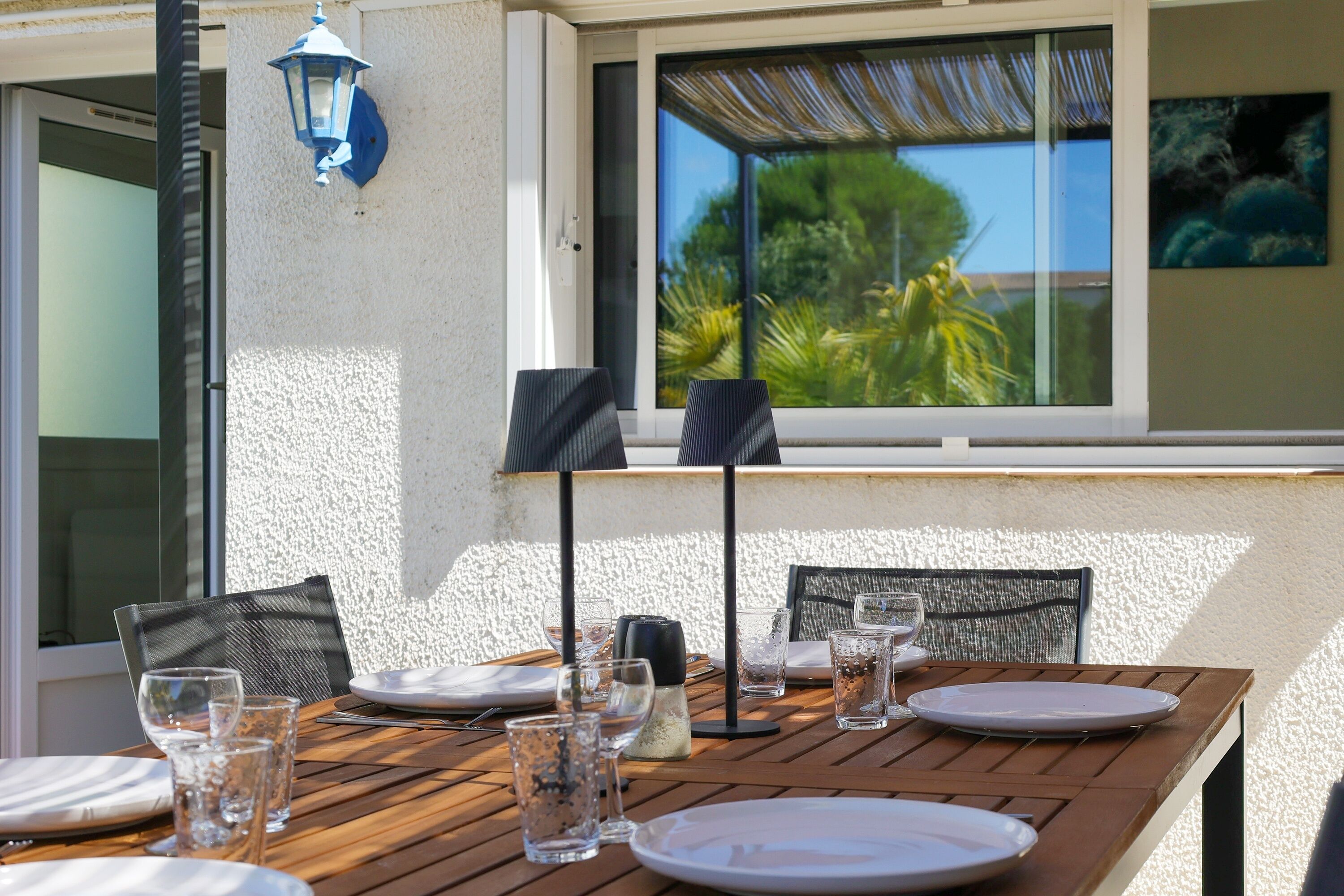 A wooden table set for dining with chairs, a window with a view of greenery, and a wall-mounted lamp.