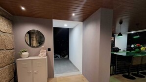 Un interior modern amb un sostre de fusta, una paret de pedra, un moble amb calaixos, un mirall rodó i una zona de bar il·luminada amb verd.