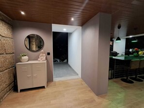 A modern interior with a wooden ceiling, a stone wall, a cabinet with drawers, a round mirror, and a green-lit bar area. - Casa de Santo António - Suite 1 (Marco de Canaveses)