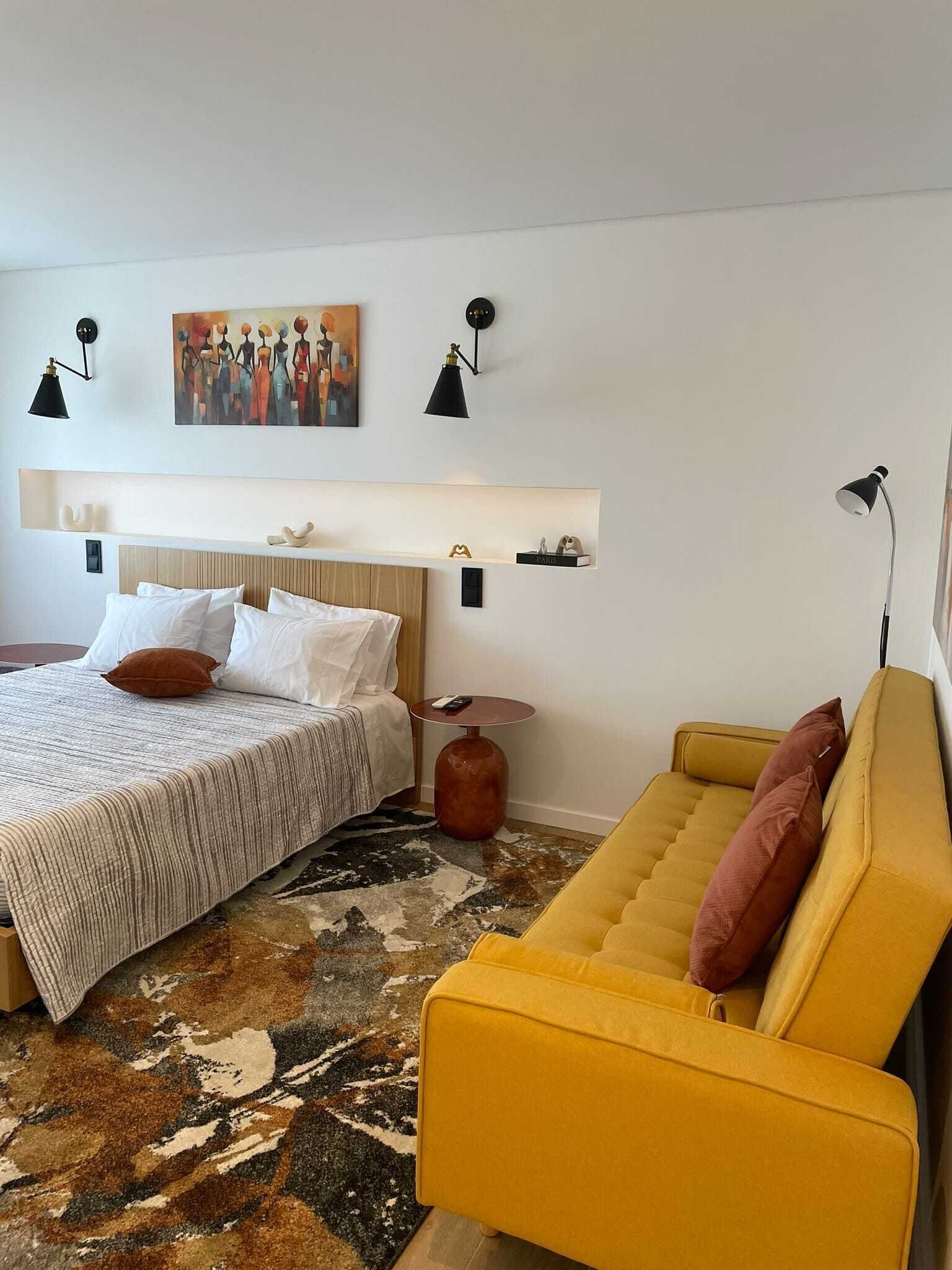 Une chambre d’hôtel avec un lit, un canapé, un tapis, un tableau et des lampes fixées au mur.
