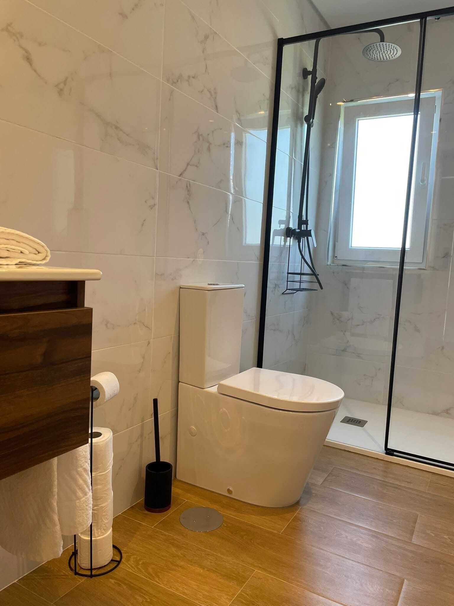 Une salle de bain comprenant des toilettes, une douche, un mur recouvert de carreaux de marbre, un meuble-lavabo en bois et un porte-rouleau de papier toilette.