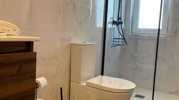 Une salle de bain comprenant des toilettes, une douche, un mur recouvert de carreaux de marbre, un meuble-lavabo en bois et un porte-rouleau de papier toilette.