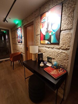 A hotel lobby with a stone wall, a large abstract painting, a wooden door, a desk with a globe, a lamp, and a chair.
