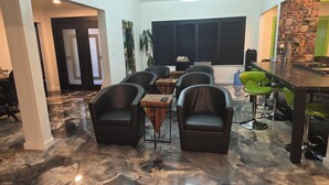 Un área de comedor moderna con piso de mármol, sillas de cuero negro, una barra con taburetes verdes y un ventanal con persianas.