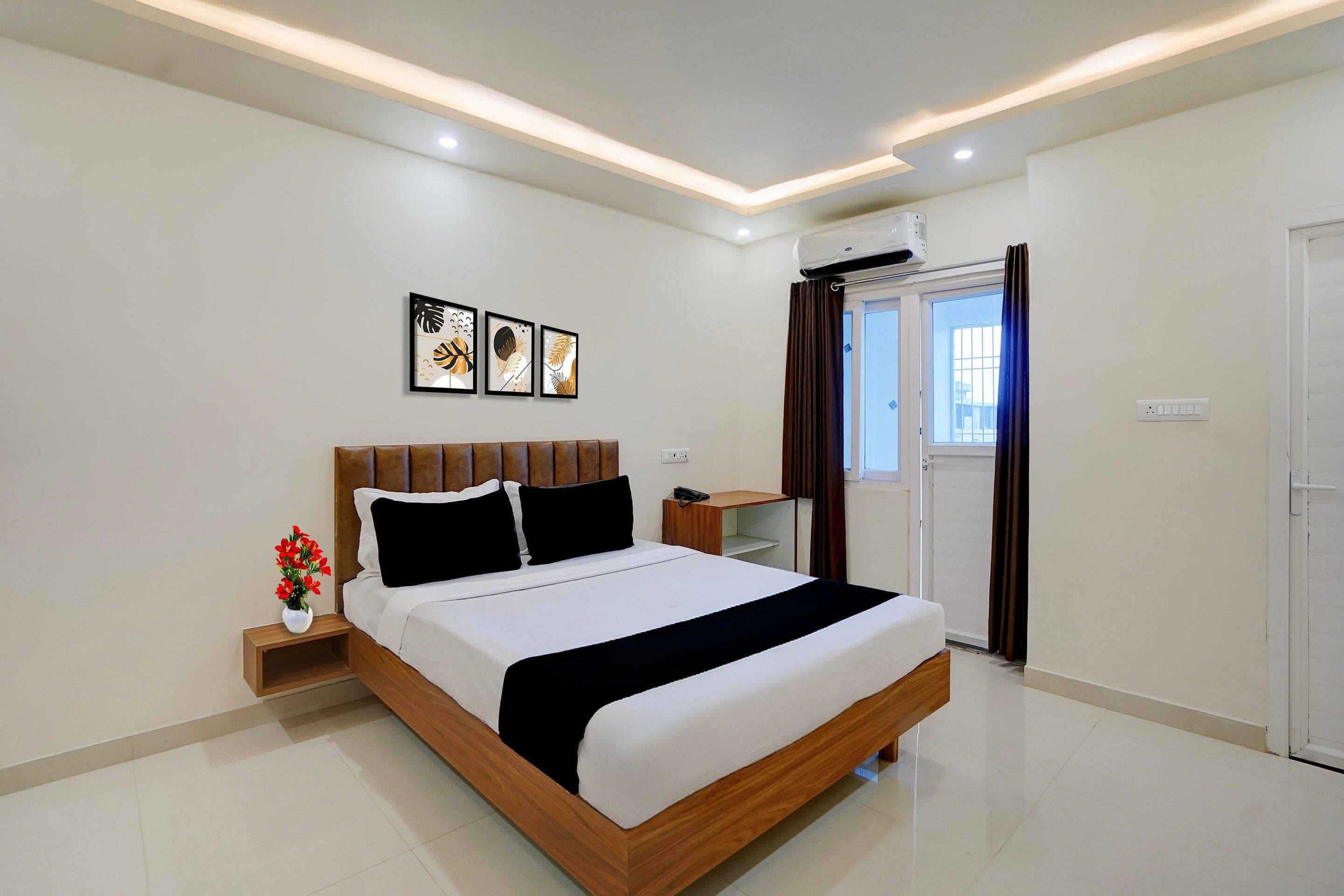 A modern hotel room with a large bed, a bedside table, a vase of flowers, and a window with curtains.
