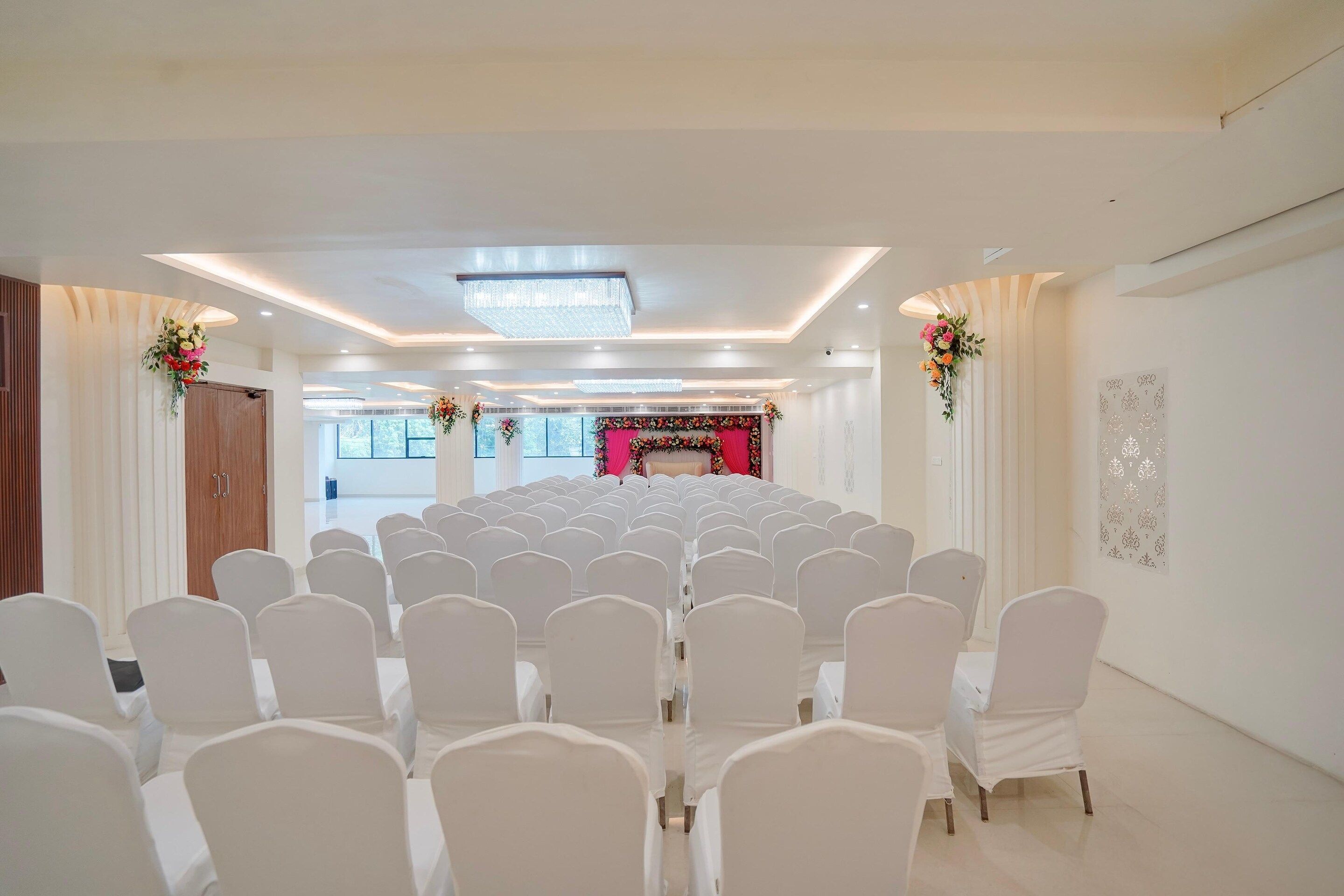 Uma sala de conferências com fileiras de cadeiras brancas, um palco com decoração floral e um design de teto moderno.