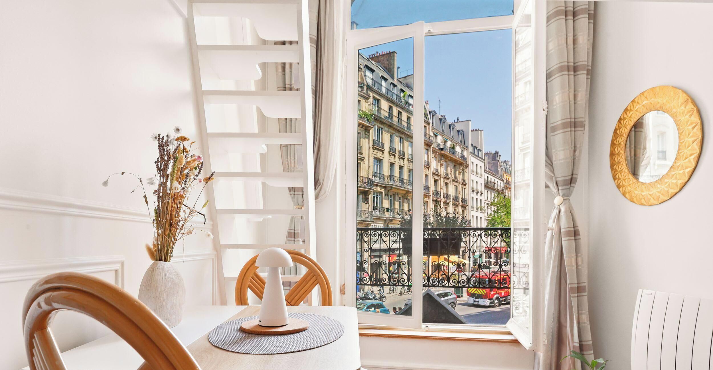 A room with a dining table, chairs, a vase with dried flowers, a window with a view of buildings, and a decorative mirror.