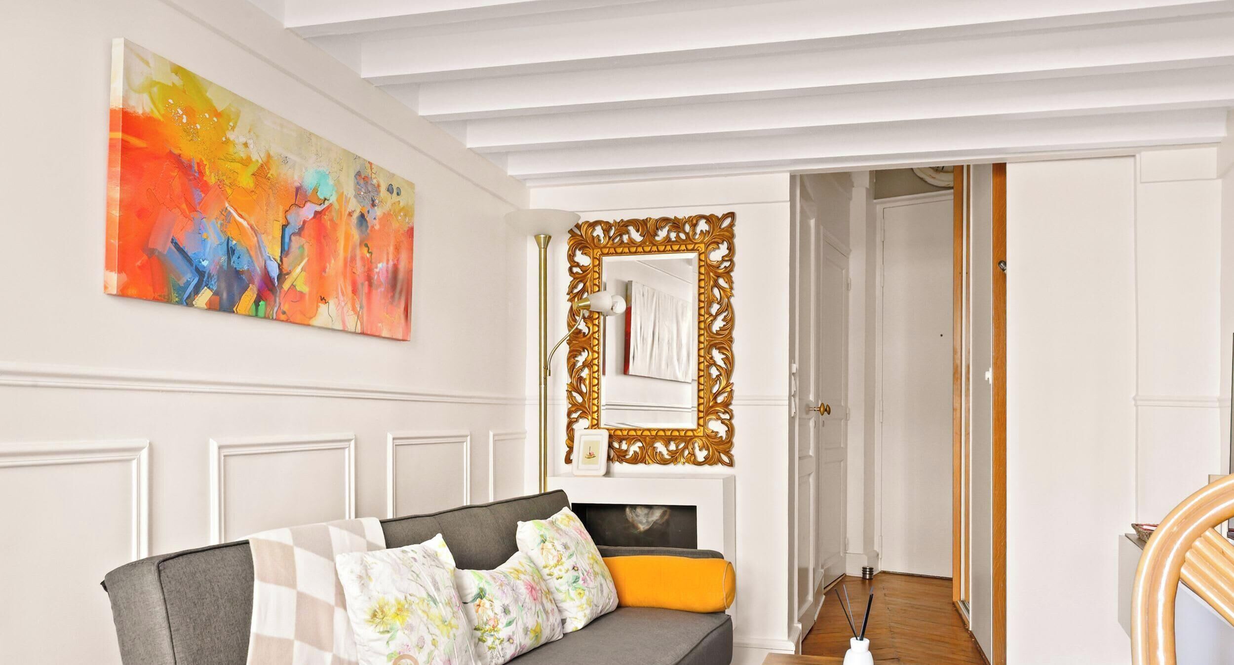 Une pièce avec un canapé gris, des coussins à motifs floraux, un grand tableau abstrait et un miroir doté d’un cadre orné.