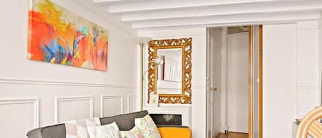 Une pièce avec un canapé gris, des coussins à motifs floraux, un grand tableau abstrait et un miroir doté d’un cadre orné.