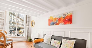 Ein Zimmer mit grauem Sofa, blumigen Kissen, einem großen abstrakten Gemälde, einem Fenster mit Blick auf Gebäude und einem Korbstuhl.