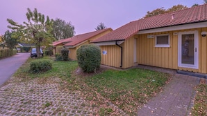A row of single-story buildings with a tiled roof, a paved driveway, and a small garden area with a hedge. - Skandinavischer Doppelbungalow mit Terrasse im Ferienpark Mirow (Mirow)